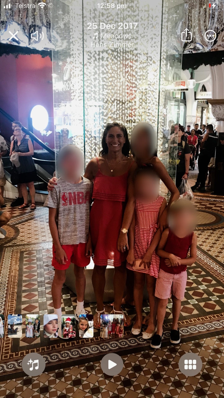 A woman smiles in a summer dress with her arms around four children whose faces are blurred. 