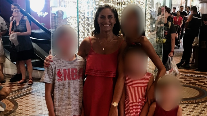 A woman smiles in a summer dress with her arms around four children whose faces are blurred. 