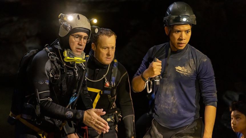 Two white men and a Thai man wear scuba diving gear and head torches in a dark cave and look intently at something off camera