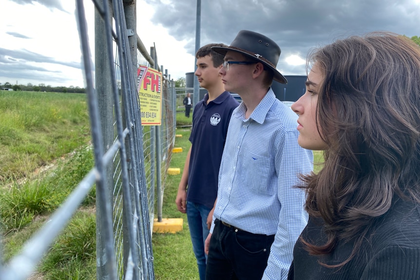 Three students stand in front of a fence watching an empty field where their school is supposed to stand.
