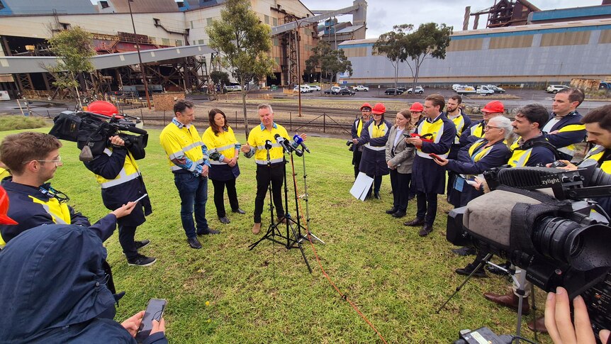 Chris Bowen addresses a large pack of media with microphones in front of him and steelworks in the background.