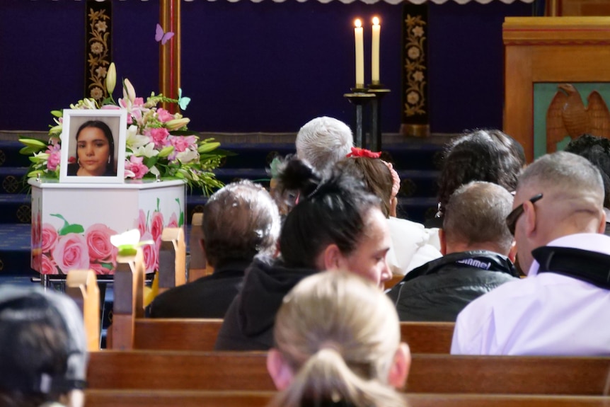 A group of people sit in church pews with a casket to the left.