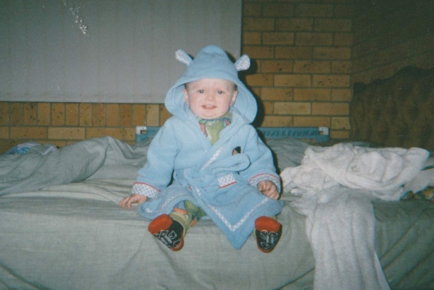 A toddler in a blue onesie ith ears, wears socks, sits on an unmade bed with a velvet brown bedhead.