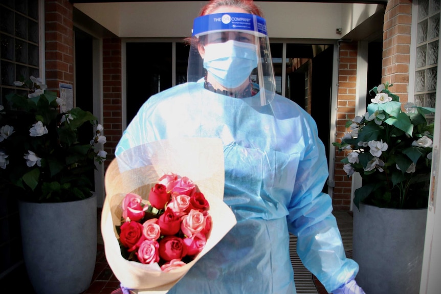 A woman wears full PPE gown, mask, shield and gloves, while holding a big bunch of flowers.