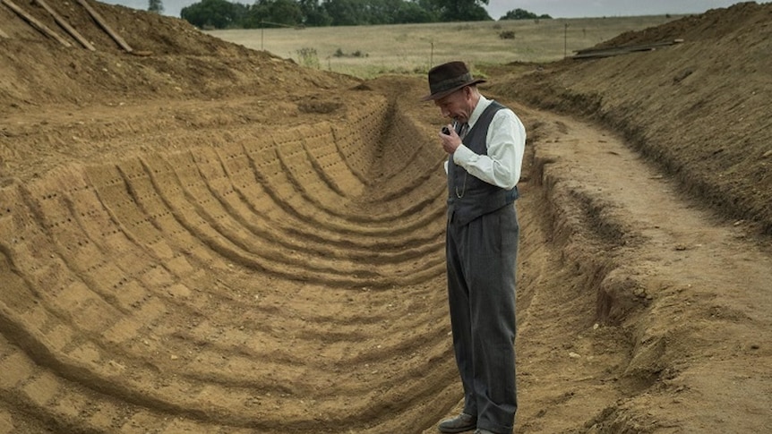 Image from The Dig (2021 movie) starring Ralph Fiennes as excavator Basil Brown looking at boat shape uncovered at Sutton Hoo.