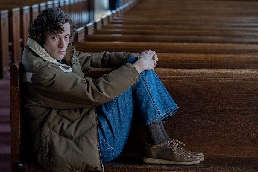 A film still of Dominic Sessa, a young man, sitting in a church pew, his feet on the seat and hands on his knees.