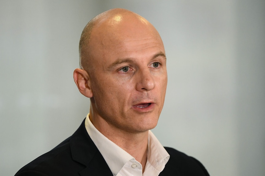 A bald man opens his mouth to speak while wearing a white shirt and black suit.