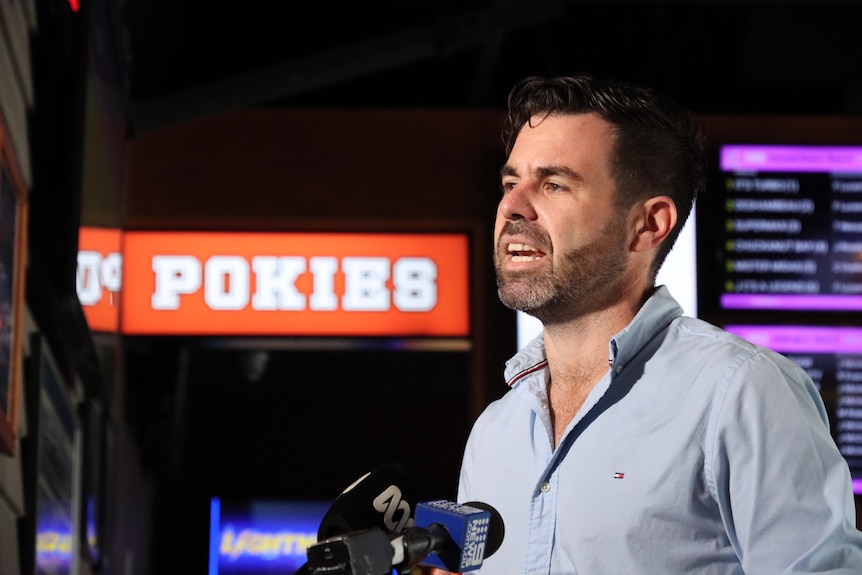A man standing in front of microphones, with pokies in the background.