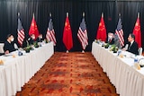 Women and men in suits sit a opposing long tables, with Chinese and US flags hanging in the background.