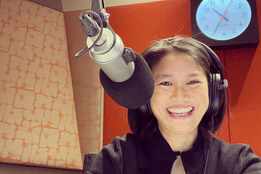 Lisa Leong, wearing headphones, with a microphone in front of her and a studio clock behind her, smiles widely.