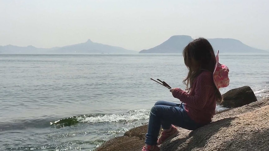 A young girls sits on a beach with islands visible in the background.