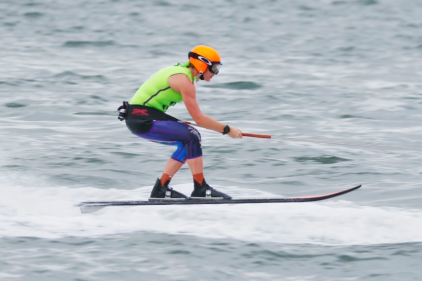 Side-on photo of a young woman waterskiing. She wears a bright coloured helmet.