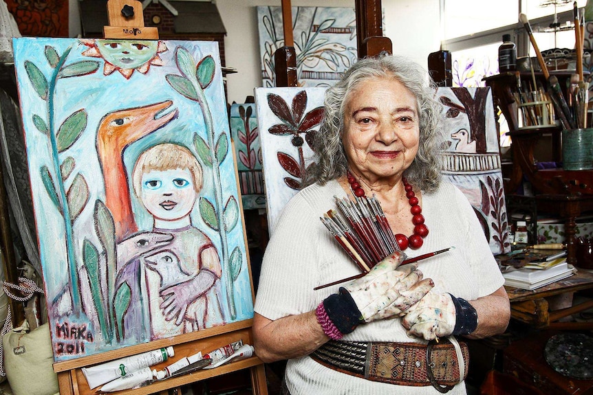 Woman wearing red beads and holding paintbrushes in her studio