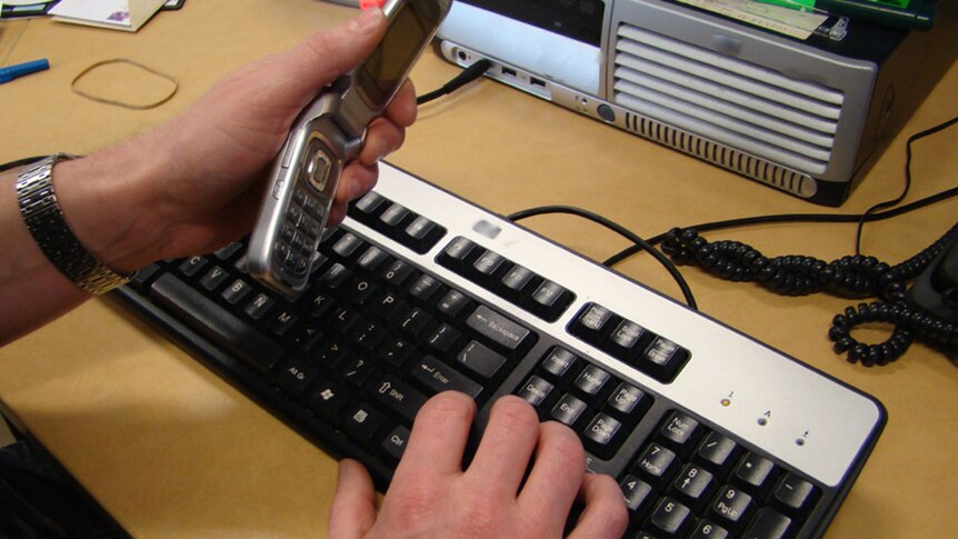 A hand holding a mobile phone and another hand hovering over a computer keyboard