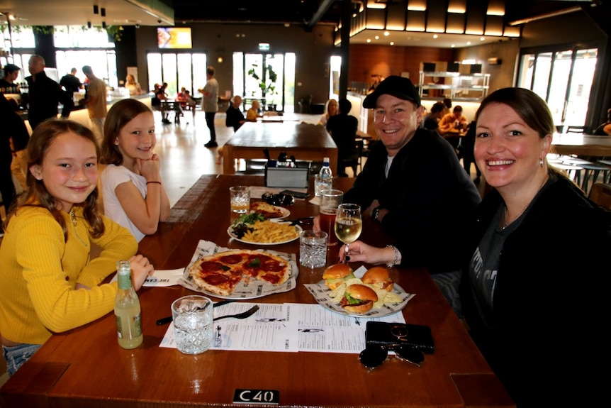 Two girls sit at a table in a pub with their parents with plates of food in front of them and patrons in the background.