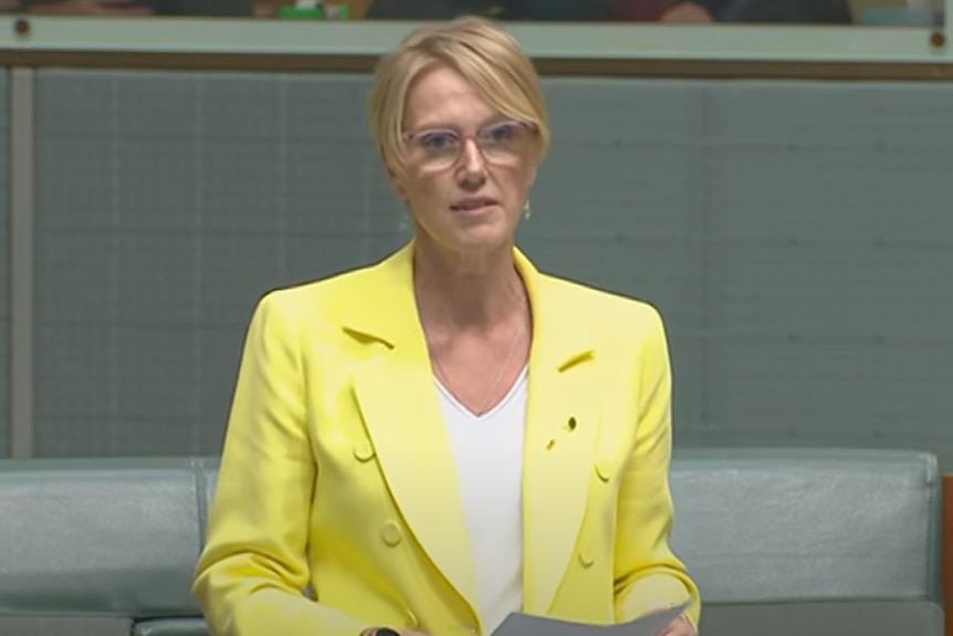A middle-aged blonde woman in glasses and a yellow jacket asks a question in a large green room.