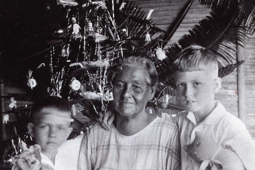 An archival, black and white photo shows a grandmother with two boys in front of a Christmas tree.