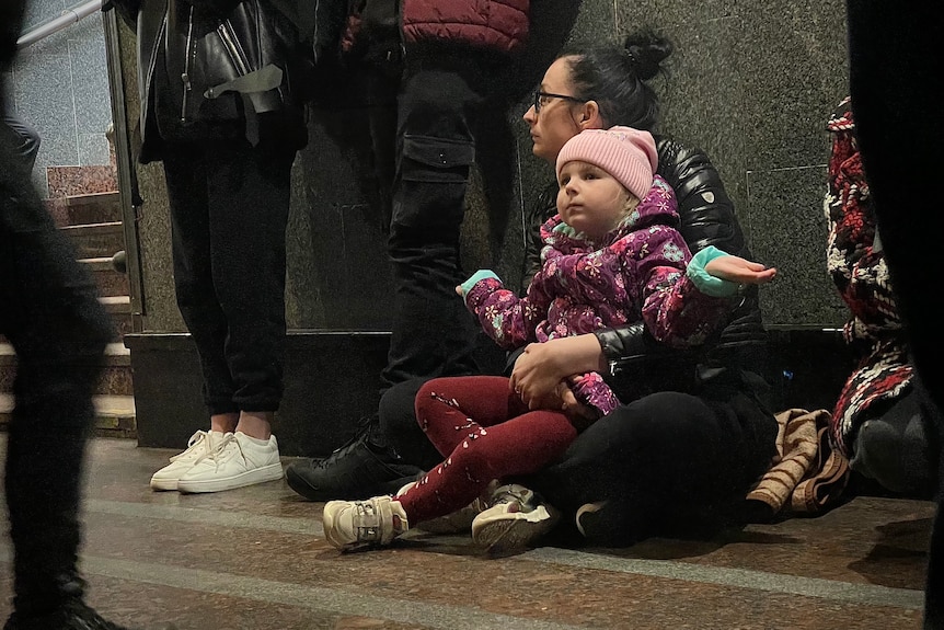 A girl sits on her mother's lap on a concrete floor. Both are wearing winter clothes