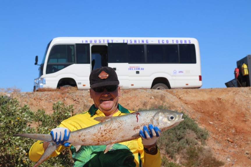 A smiling man, holding a fish, with a tourist bus in the background. 