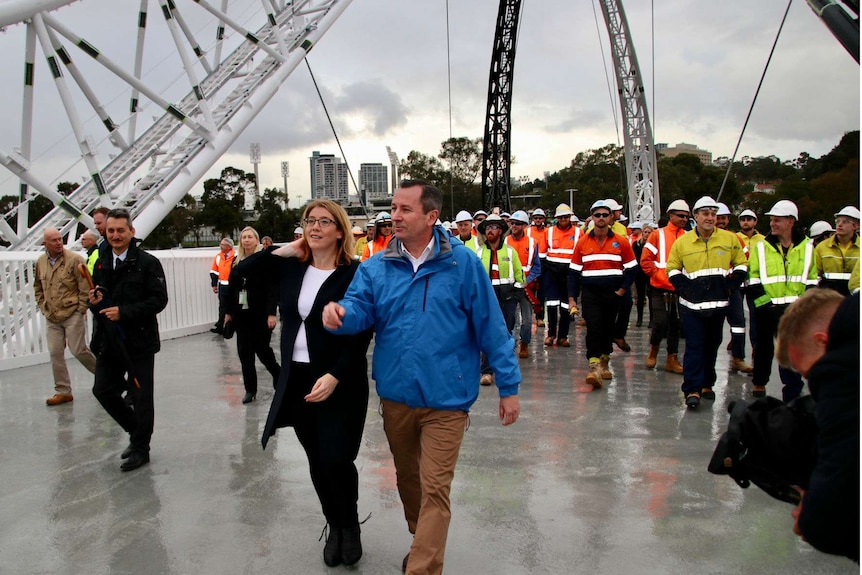 A man and a woman walk across a bridge with workers in high-vis gear behind them.