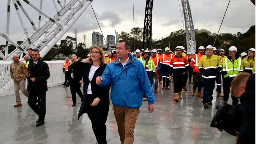 A man and a woman walk across a bridge with workers in high-vis gear behind them.