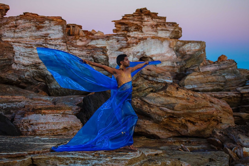 Indigenous man with blue silk wings spread, standing in front of red rocks and sunset