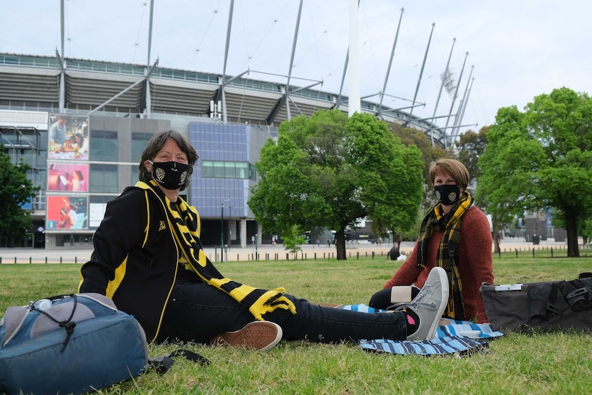 Two women sit in the grass wearing Richmond face masks with the MCG in the background.
