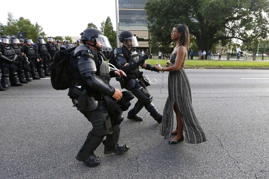 A woman in a dress stands neutrally as two police in armour rush in to detain her