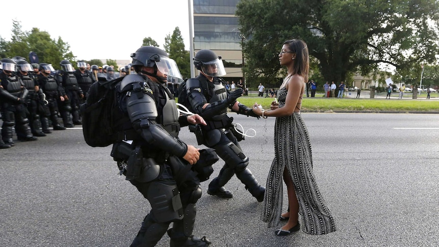 A woman in a dress stands neutrally as two police in armour rush in to detain her
