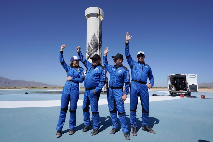 four people in blue space suits with hands in the air waving standing on a stage