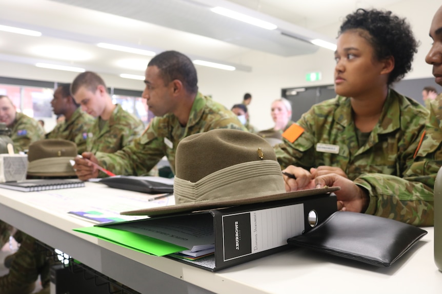 An army hat sits on a desk with books and students in the background