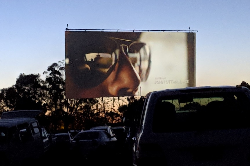 A large screen is shown above several rows of cars at a drive-in theatre. The sun is setting behind trees in the background.