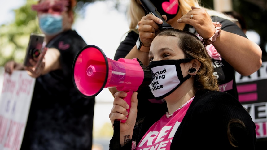 A woman wearing a white mask appears to speak into a pink megaphone while another woman shaves her head