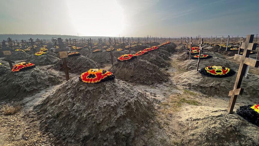 Rows and rows of graves are lined up in a dirt field, with crosses and floral arrangements adorning each one