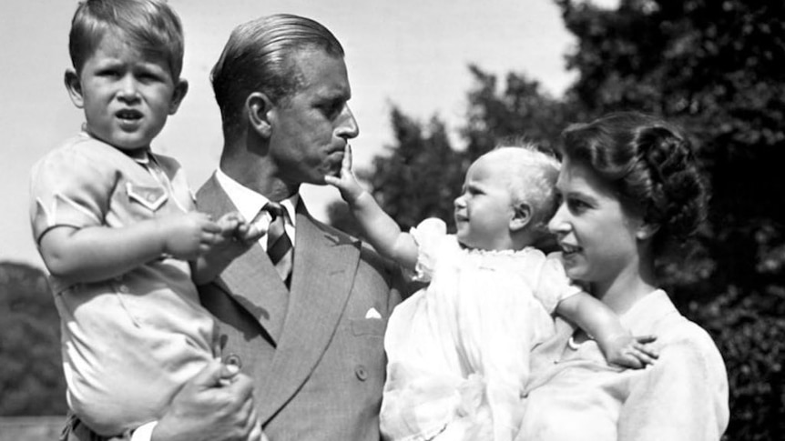 A black and white photo of a man holding a young boy and a woman holding a baby that is touching the man's mouth.