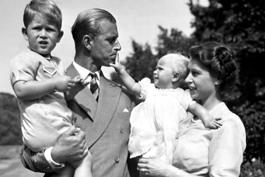 A black and white photo of Prince Philip holding boy Charles and Princess Elizabeth holding baby Anne.