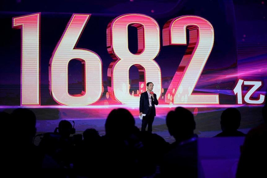 A man stands on stage in front of a giant sign read "1682".