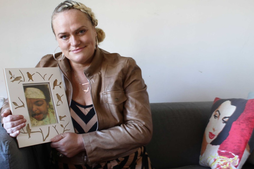 Tansy Mayhew sitting on her couch, holding a portrait of her daughter, Chanté.