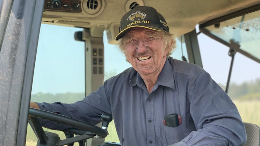 A man in a blue shirt and baseball cap leans on the wheel while sitting in the cab of a tractor.