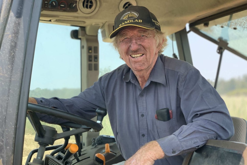 A man in a blue shirt and baseball cap leans on the wheel while sitting in the cab of a tractor.