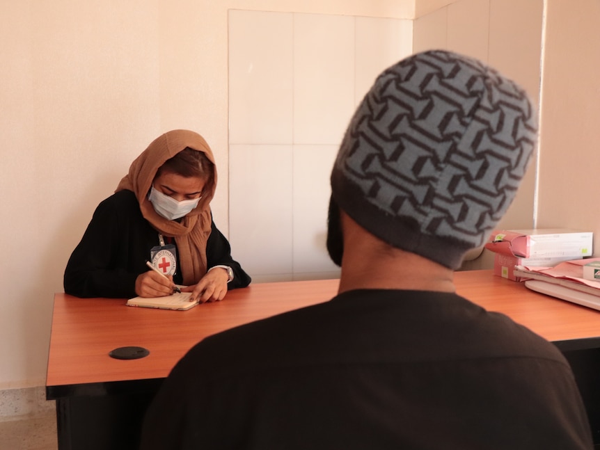 A woman in a head scarf taking notes while she speaks to a man across the table from her who's back is to the camera