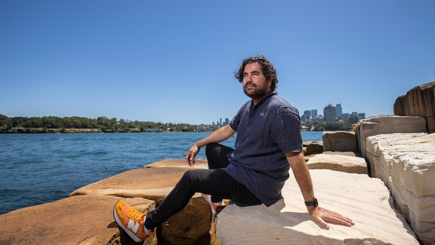 Indigenous man with curly dark hair and beard wears a navy t-shirt and sits on sandstone blocks looking over ocean.