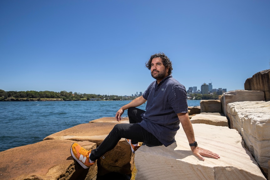 Indigenous man with curly dark hair and beard wears a navy t-shirt and sits on sandstone blocks looking over ocean.