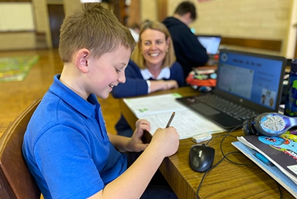 A student works at a desk as a teacher kneels alongside and smiles.