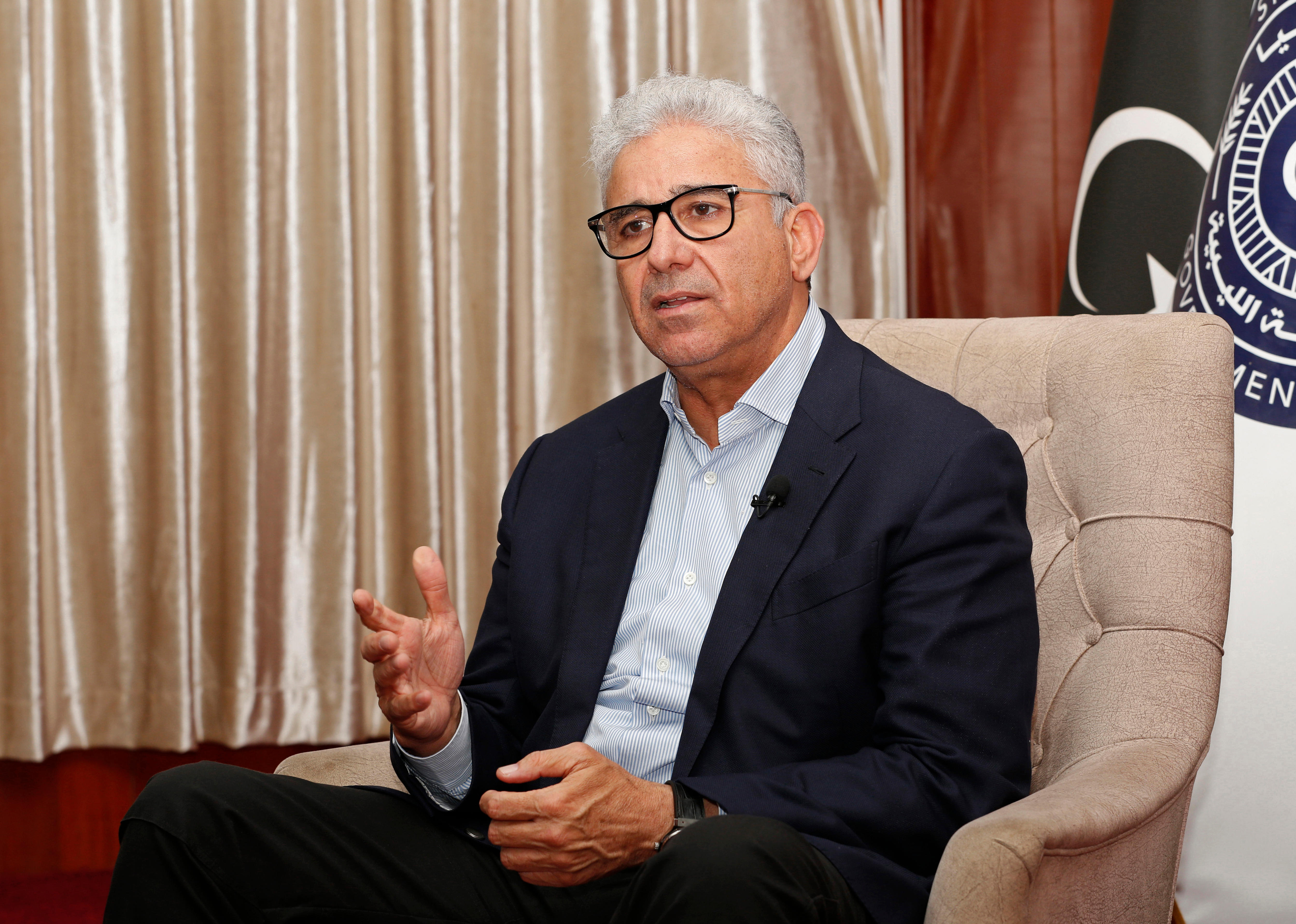 An older man with glasses and grey hair is pictured wearing a suit without a tie. 