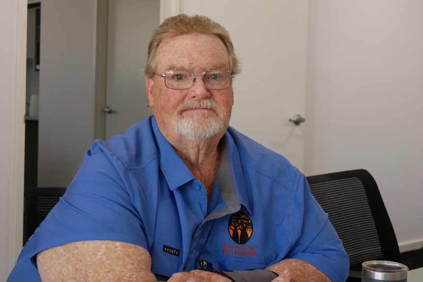 Man in blue shirt, glasses and white short beard sitting on chair looking at camera.