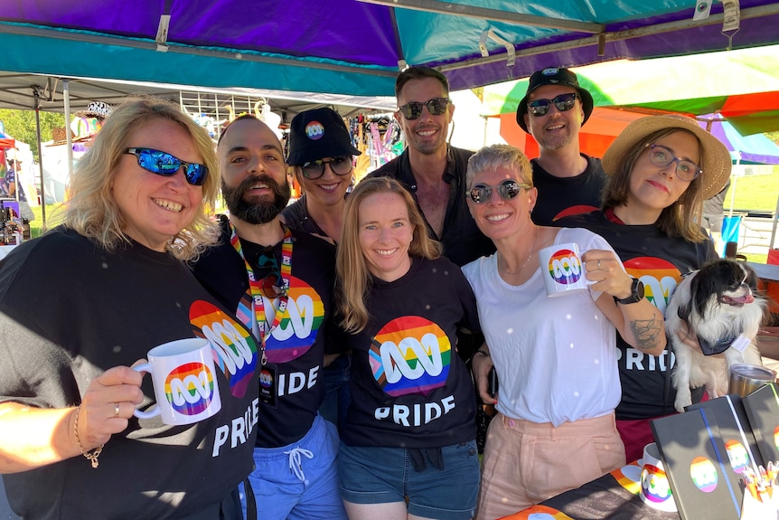 Eight people all smiling and wearing tshirts with the ABC lissajou on a background of the LGBTQIA+ pride rainbow.