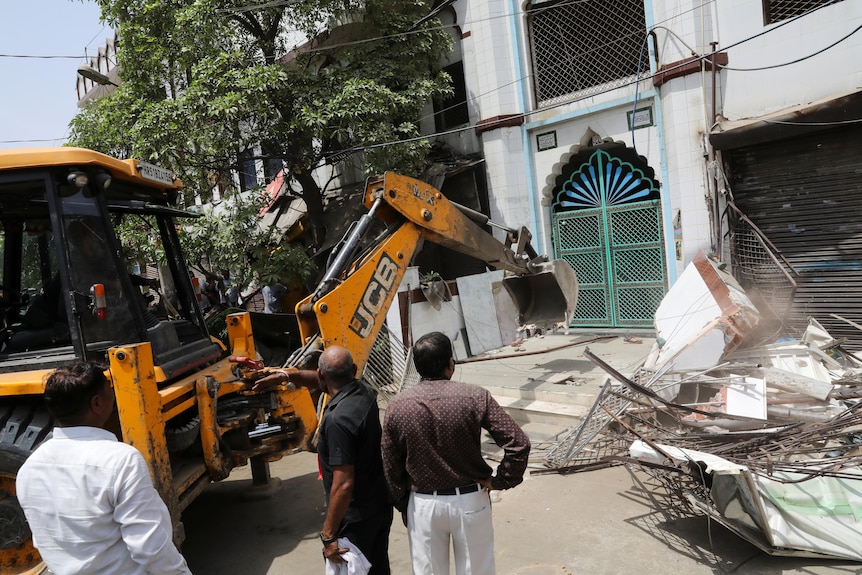 People look on as an excavator approaches a turquoise gate.