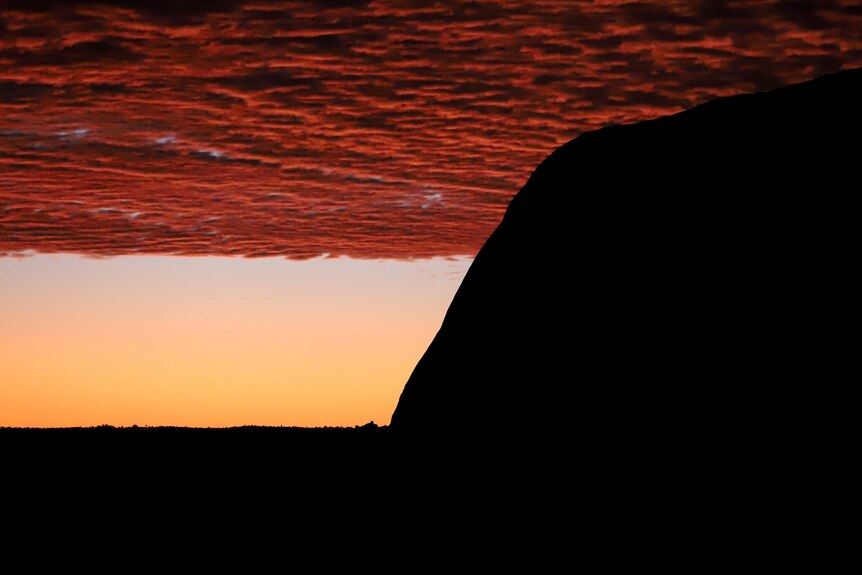 A silhouette of Uluru with reddish yellow sky and clouds