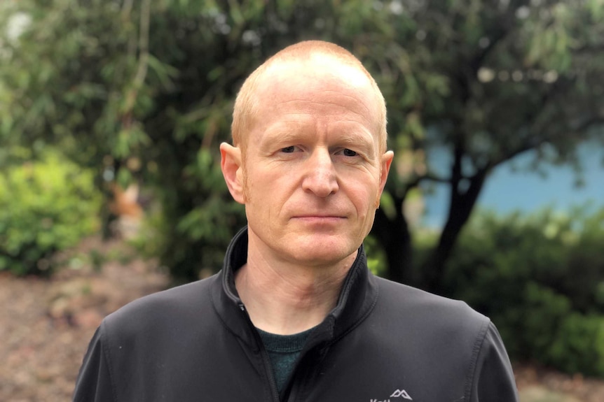 A man with short red hair with a neutral expression wearing a black jumper stares at the camera.